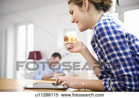 Stock Photography - Young woman in blue checked shirt eating an apple and using a tablet. Fotosearch