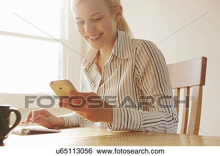 Young woman sitting at table looking at smartphone View Large Photo Image Stock Photograph - Young woman sitting at table looking at smartphone. Fotosearch