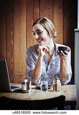 Young woman sitting in cafe holding hot drink, using laptop computer View Large Photo Image Stock Image - Young woman sitting in cafe holding hot drink, using laptop computer. Fotosearch