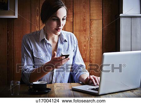 Picture - Young woman sitting in cafe holding smartphone, using laptop computer. Fotosearch