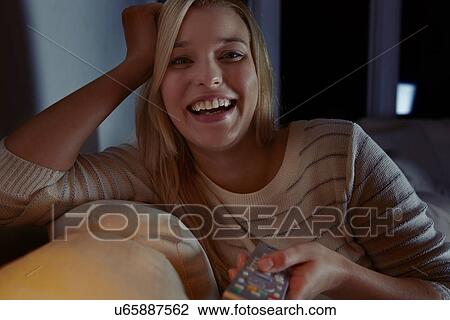 Stock Image - Young woman sitting on sofa, holding remote control. Fotosearch