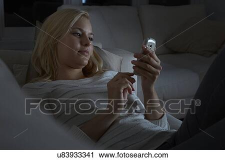 Stock Image - Young woman sitting on sofa, using smartphone. Fotosearch