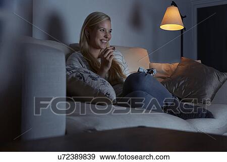 Stock Photo - Young woman sitting on sofa, watching tv, eating chocolate. Fotosearch
