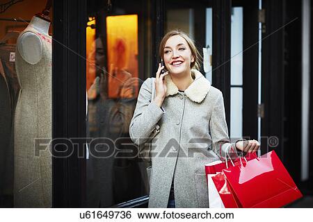 Stock Photograph - Young woman walking down street carrying shopping bag and making phone call. Fotosearch