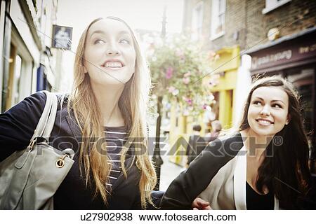 Stock Photo - Young women walking down street carrying bags and laughing. Fotosearch