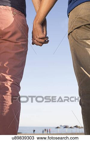Stock Image - Cropped shot of mature male couple holding hands on beach. Fotosearch