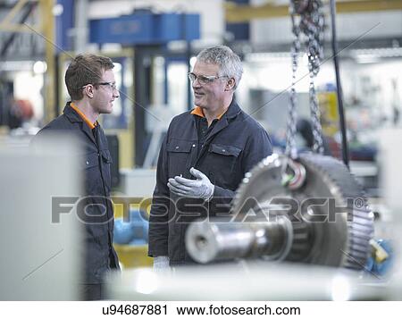 Stock Image - Engineer and apprentice in discussion next to gear wheel at work station in factory. Fotosearch