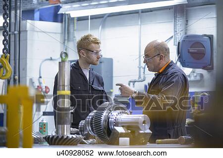 Stock Image - Engineer instructing apprentice at workstation in factory. Fotosearch