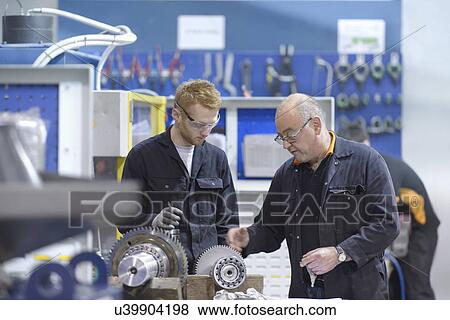 Stock Photo - Engineer instructing apprentice at workstation in factory. Fotosearch