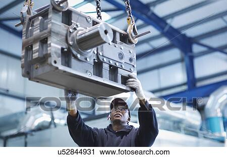 Stock Image - Engineer using crane to move industrial gearbox to paint works in engineering factory. Fotosearch