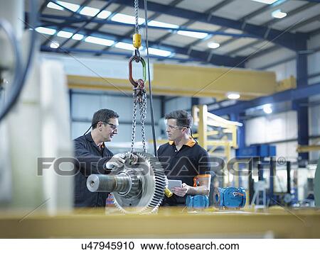 Stock Image - Engineers with gear wheel at work station in factory. Fotosearch