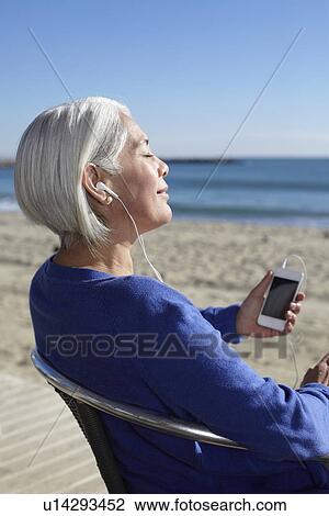 Stock Image - Mature woman listening to music with earbuds on beach. Fotosearch
