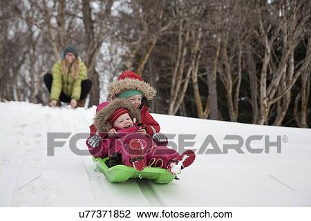 Stock Image - Mother watching two young daughters sledging in snow. Fotosearch