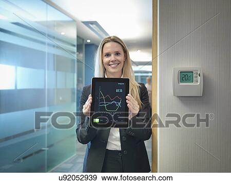 Stock Photo - Portrait of office worker holding digital tablet next to office thermostat adjusted to make energy savings. Fotosearch