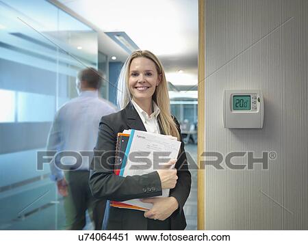 Stock Image - Portrait of office worker next to office thermostat adjusted to make energy savings. Fotosearch