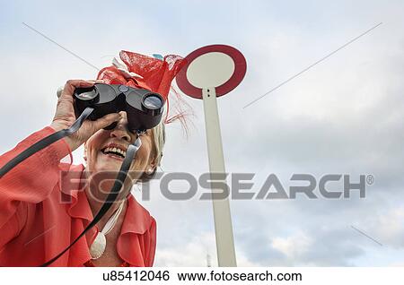 Stock Photograph - Senior woman at races leaning forward and looking through binoculars. Fotosearch