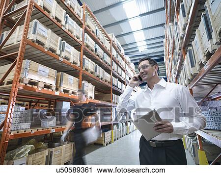 Stock Image - Worker using mobile phone and holding digital tablet in parts store in factory. Fotosearch