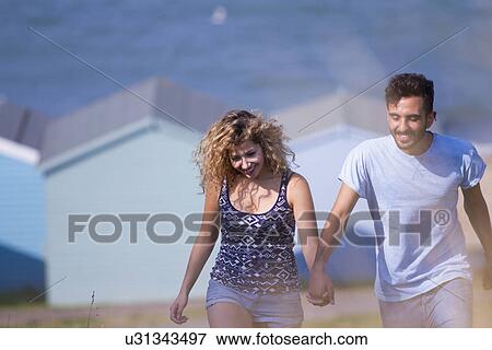 Stock Photo - Young couple holding hands with beach huts in background. Fotosearch