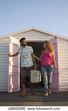 Stock Image - Young couple leaving beach hut with suitcase. Fotosearch