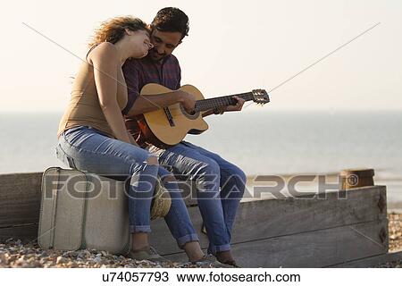 Stock Image - Young couple on beach, man playing guitar. Fotosearch