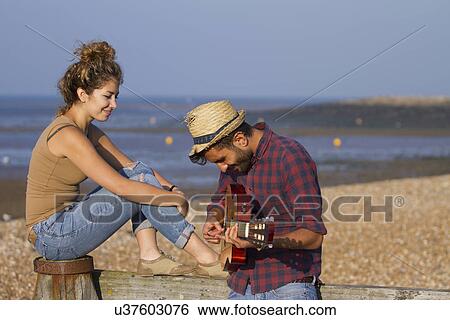 Stock Photograph - Young couple on beach, man playing guitar. Fotosearch