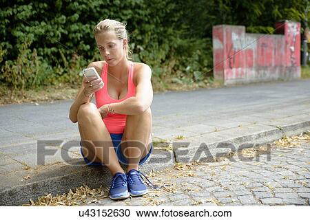 Stock Image - Young female runner sitting on sidewalk using cellphone. Fotosearch