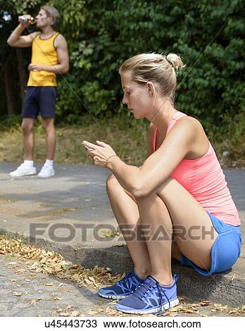 Stock Image - Young female runner sitting on sidewalk looking at cellphone. Fotosearch