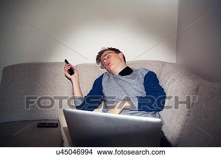 Picture - Young man asleep on sofa with remote control and laptop. Fotosearch