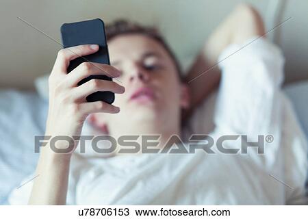 Young man lying in bed looking at mobile phone View Large Photo Image Stock Image - Young man lying in bed looking at mobile phone. Fotosearch