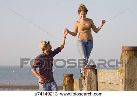 Stock Image - Young woman balancing on groynes holding man's hand. Fotosearch