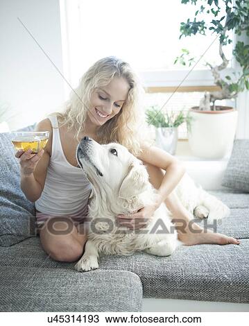 Stock Image - Beautiful woman holding tea cup while touching dog on sofa. Fotosearch