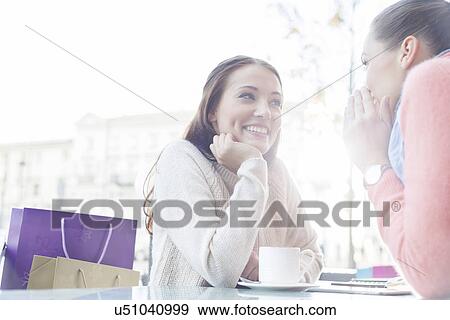Stock Photo - Beautiful young woman looking at friend sharing secrets at outdoor cafe. Fotosearch