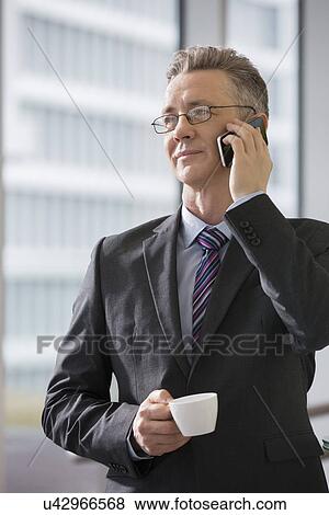Stock Photo - Businessman with coffee cup using cell phone in office. Fotosearch