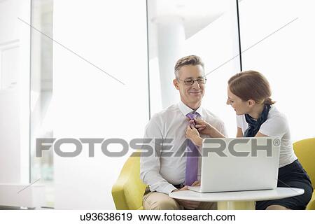 Stock Photo - Businesswoman putting on tie to male colleague at lobby. Fotosearch