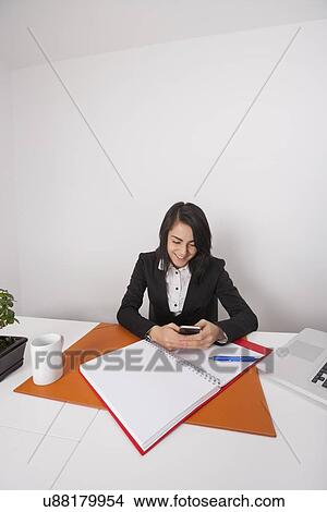 Businesswoman text messaging through cell phone at office desk View Large Photo Image Picture - Businesswoman text messaging through cell phone at office desk. Fotosearch