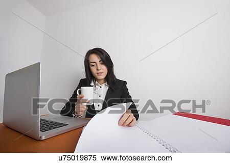 Stock Photography - Businesswoman with coffee mug reading documents at desk in office. Fotosearch