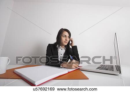 Businesswomen using cell phone while writing notes from laptop at office desk View Large Photo Image Stock Photo - Businesswomen using cell phone while writing notes from laptop at office desk. Fotosearch