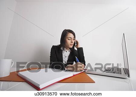 Stock Image - Businesswomen writing notes while using cell phone at office desk. Fotosearch