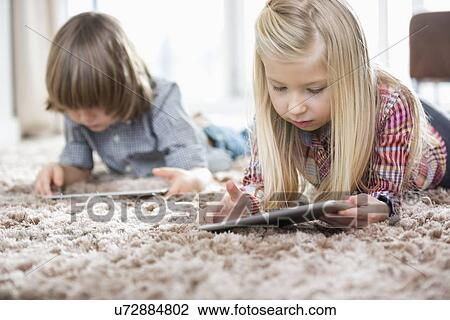 Stock Image - Cute girl and brother using digital tablets on rug in living room. Fotosearch