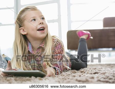 Cute girl with digital tablet looking away while lying on rug in living room View Large Photo Image Stock Photograph - Cute girl with digital tablet looking away while lying on rug in living room. Fotosearch