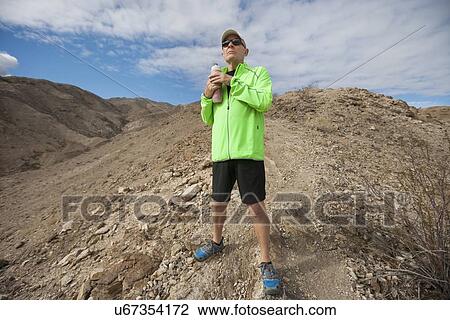 Stock Image - Determined senior man holding water bottle while standing on mountain. Fotosearch