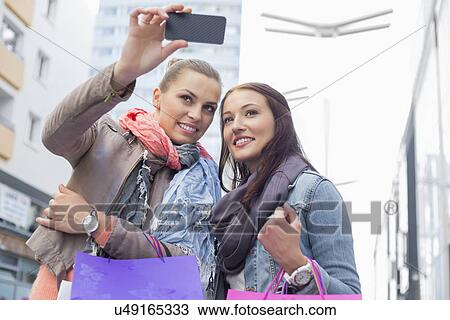 Stock Image - Female friends with shopping bags taking photos through mobile phone. Fotosearch