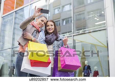 Female friends with shopping bags taking self portrait through mobile phone against store View Large Photo Image Stock Photo - Female friends with shopping bags taking self portrait through mobile phone against store. Fotosearch