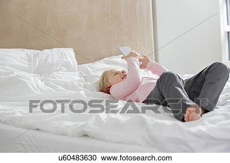 Stock Image - Full length of happy girl using smart phone in bed. Fotosearch
