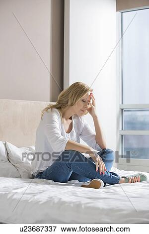 Stock Photo - Full length of stressed woman with hand on face sitting on bed. Fotosearch