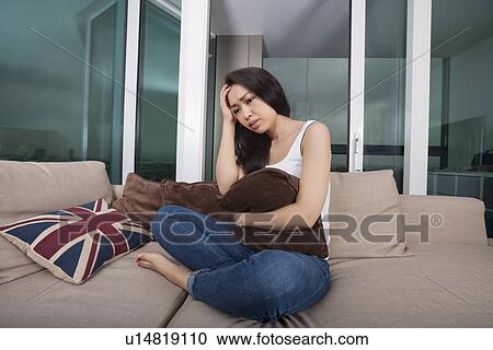 Stock Image - Full length of worried young woman sitting on sofa. Fotosearch