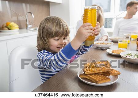 Stock Photograph - Happy boy pouring honey on waffles while having breakfast with family. Fotosearch