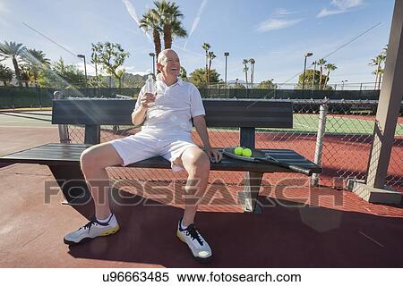 Stock Photography - Happy senior male tennis player with water bottle sitting on bench at court. Fotosearch