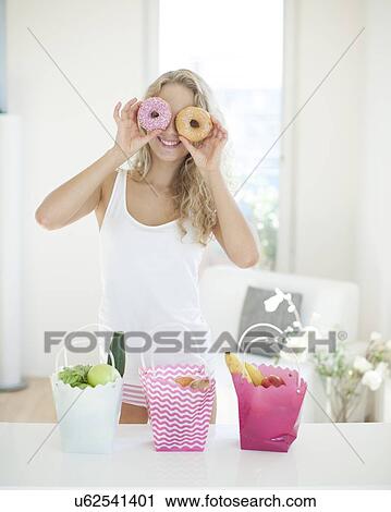 Stock Image - Happy woman holding donuts in front of eyes at kitchen counter. Fotosearch