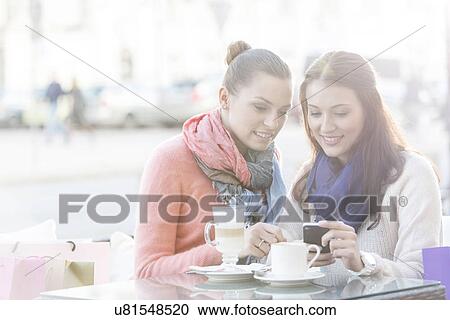 Stock Image - Happy women using cell phone at sidewalk cafe during winter. Fotosearch